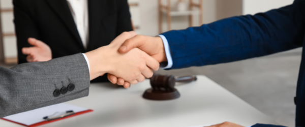 Two individuals in professional attire shake hands across a table with a gavel and legal documents in the foreground.