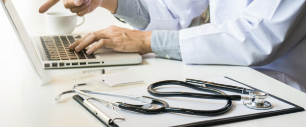 Close-up of a medical professional wearing a white coat using a laptop with a stethoscope, pen, and medical chart on a white desk.