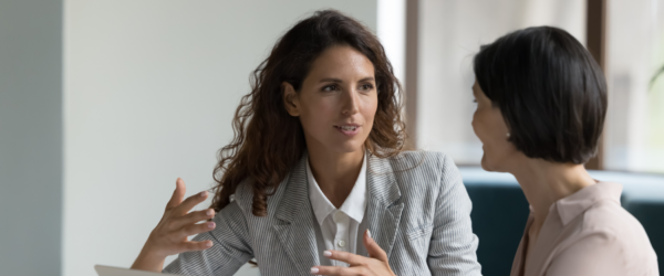 Two professionals in a business meeting, with one woman in a striped blazer gesturing while speaking to another woman.